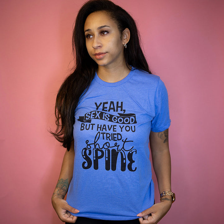 Woman in front of a pink background wearing a heather columbia blue unisex crew neck t-shirt that says "yeah, sex is good but have you tried short spine?" in black text