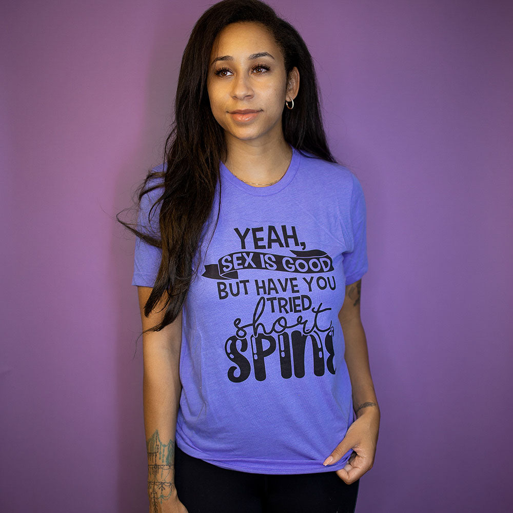 Woman in front of a purple background wearing a heather columbia blue unisex crew neck t-shirt that says "yeah, sex is good but have you tried short spine?" in black text