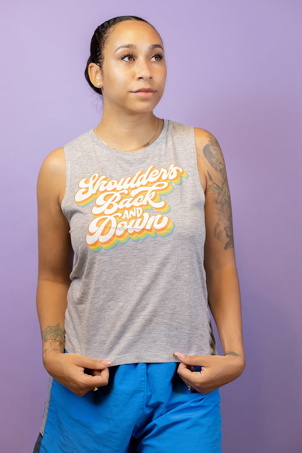Woman wearing a grey muscle tank top that says "shoulders back and down" in multi color retro script
