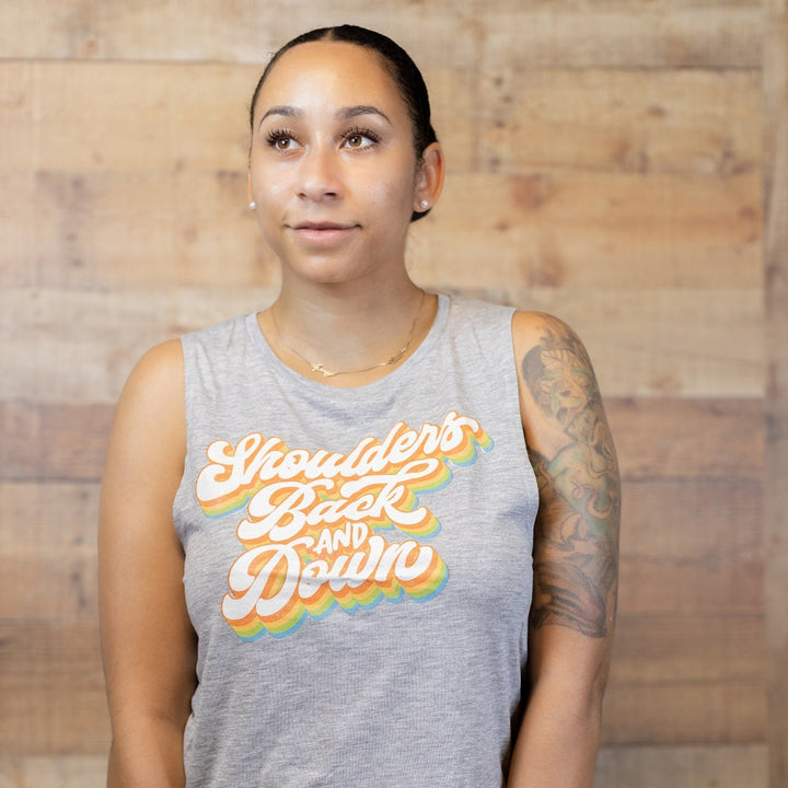 Woman wearing a grey muscle tank top that says "shoulders back and down" in multi color retro script