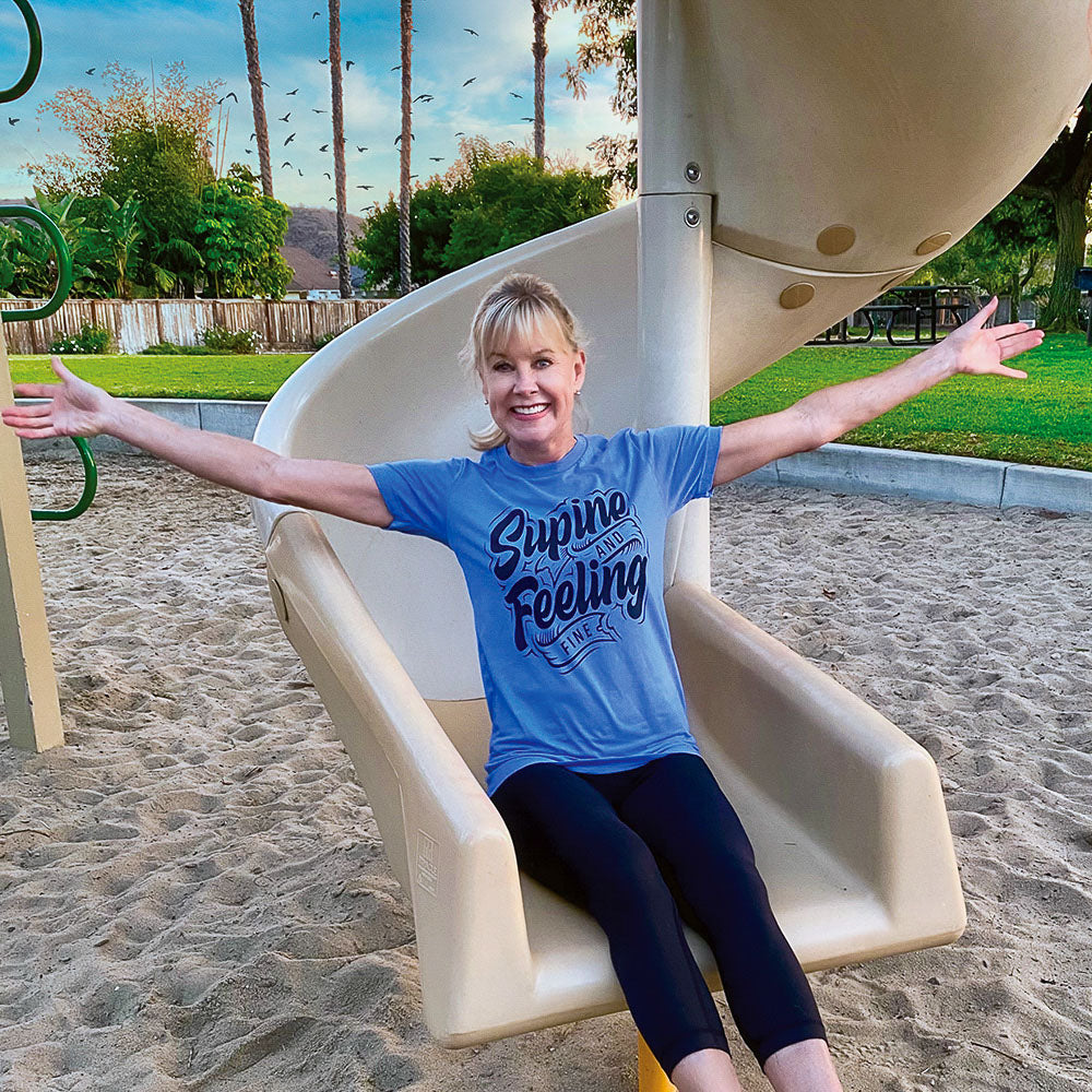 Woman wearing a blue t-shirt that says "supine and feeling fine" in black text