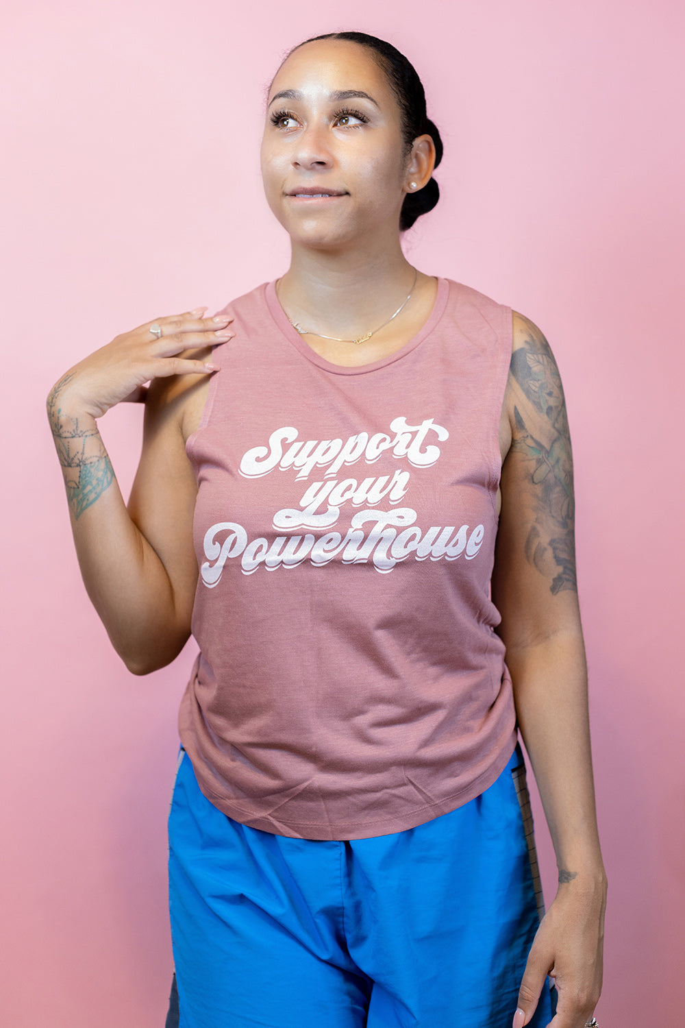 Woman standing against a pink  background wearing a mauve tank top that says "support your powerhouse"