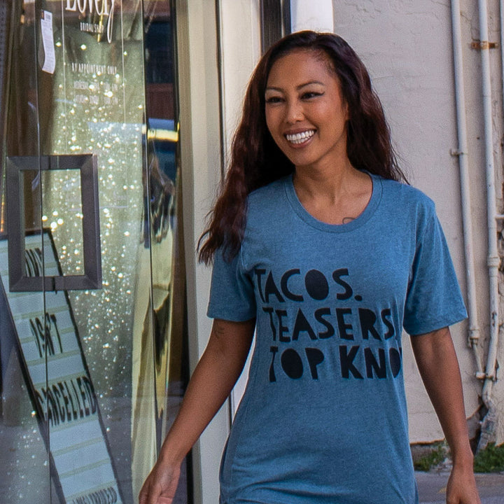 Woman wearing a blue shirt that says "Tacos. Teasers. Top Knots." in black writing