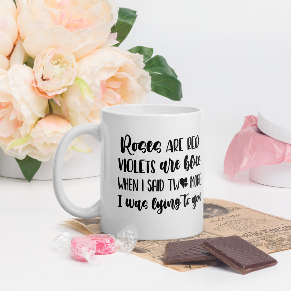 11oz white coffee mug that says "Roses Are Red, Violets Are Blue, When I Said Two More, I Was Lying To You". There are flowers in the background