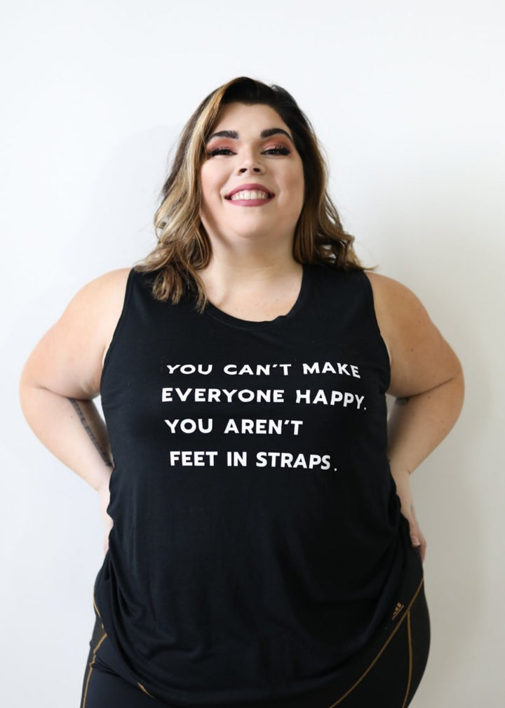 A  woman wearing a black muscle tank top that says "you can't make everyone happy; you aren't feet in straps" in white text on the front