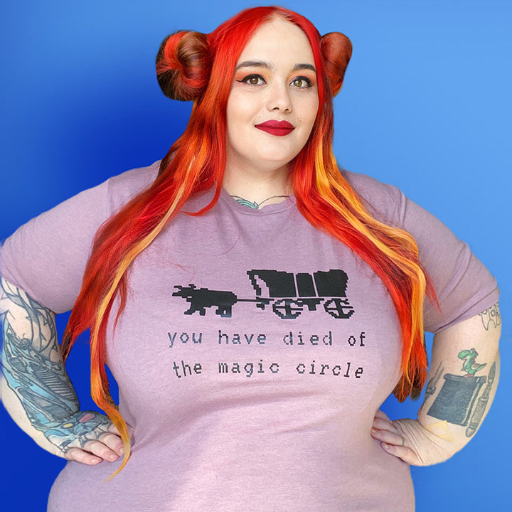 Woman wearing a unisex crewneck heather orchid t-shirt that says "You Have Died Of The Magic Circle" in 80's throwback font with the Oregon trail  ox and cart. Woman is leaning against blue background